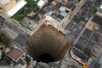 Arial photo of a large sink hole. The hole has sunk over a city block, including roads and buildings.