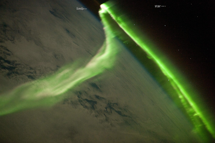 The aurora has a sinuous ribbon shape that separates into discrete spots near the lower right corner of the image. While the dominant coloration of the aurora is green, there are faint suggestions of red left of image center. Dense cloud cover is dimly visible below the aurora.