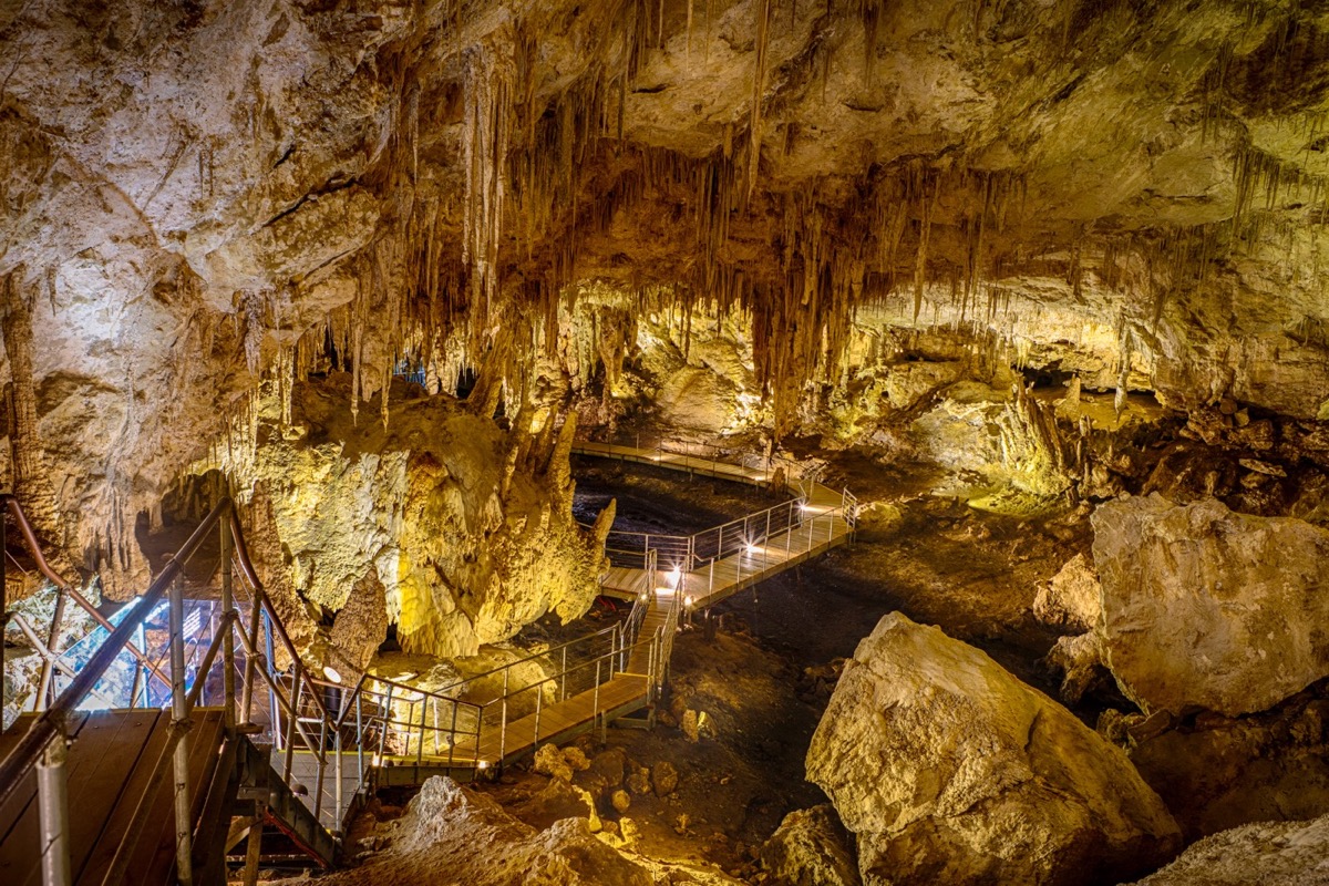 A large cave interior in golden light with stalagmites and stalagtites, and a walkway with a railing going through