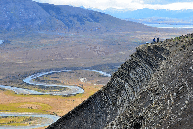 A scene in steep mountain terrain, with a meandering river in the background. In the foreground, a mountain slope reveals a succession of sedimentary layers - black shales with thin white sandstones at the base of the slope, and thicker, blockier layers of sandstone (with thin shale interbeds) at the top of the slope. At the very top stand two small geologists.