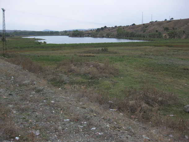 Photograph showing a pond in a low valley. Some communication towers are visible on the far side.