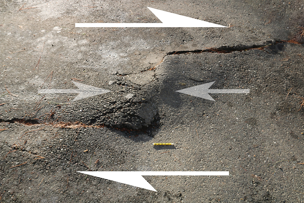 Annotated photograph of a 3m x 2m area of parking lot pavement. There are two cracks in the pavement: one coming in at the left edge of the frame, about 2/5ths of the way up from the bottom of the photo to the top, and dying out midway across the photo. A second crack starts at the right side of the photo, about 1/5th of the way down from top to bottom, and comes across to the mid-point. Between them is a bulge that is popping upward due to compression. A pencil provides a sense of scale.