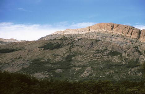Landscape photo of a region in Argentina, showing low-lying hills with vertical bedding, and they are topped by big cliffs of volcanic layers.