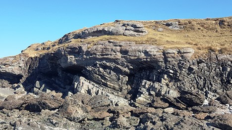 Photograph showing a Scottish seaside cliff outcrop, with thin right-dipping layers below, and horizontal, thick layers above.