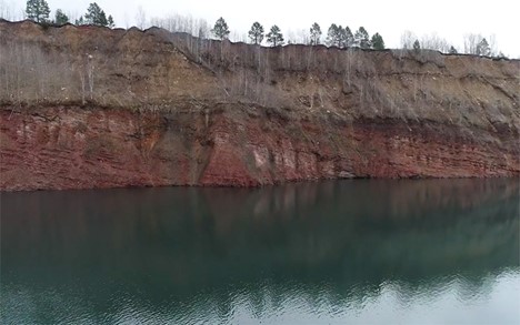 Photograph of a cliff next to a lake. The lower half of the cliff is a steep wall of red rock layers, gently dipping off to the right. Above that is a grayish zone that has more trees and appears to weather more easily.