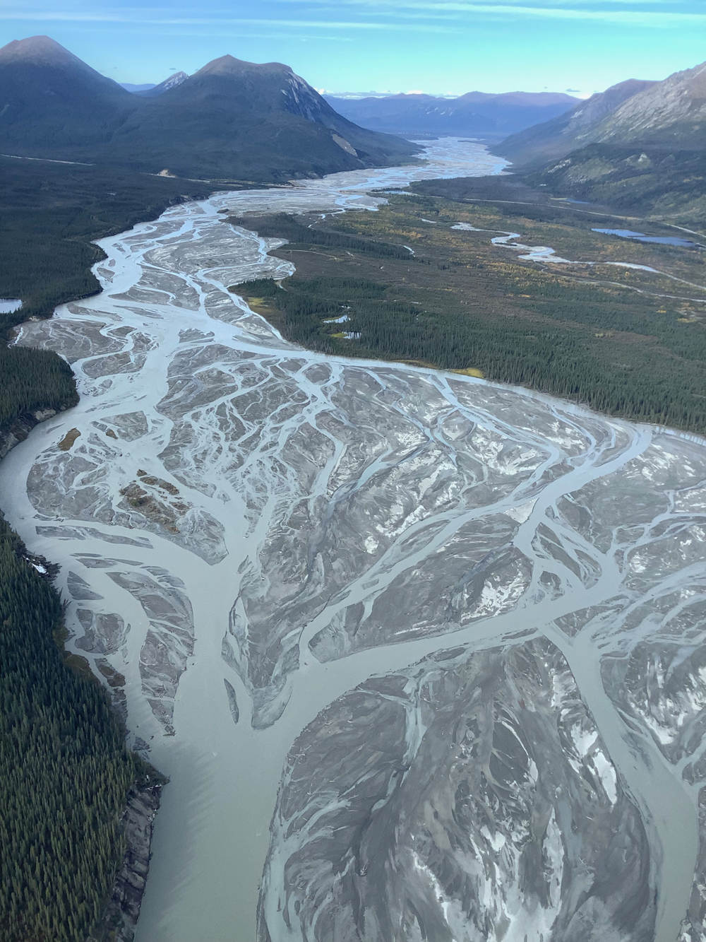 Aerial photograph of the Donjek River, a modern braided river, Yukon Territory, Canada. A wide river valley between mountain ranges is shown, with about half the valley's width covered by a vast, shiny fresh deposit of gray gravel sediment. Countless branching and re-merging channels are full of gray/blue suspended sediment. The remainder of the valley has scrubby forest covering it. The mountain slopes are bare - suggesting a very cold climate.
