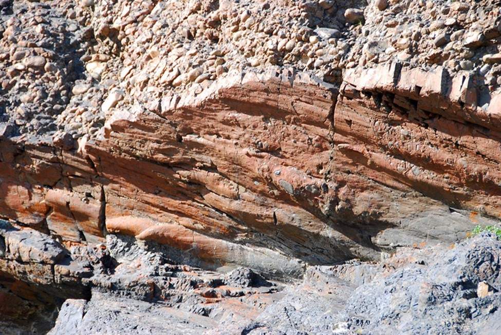 Photograph showing an oblique view of the contact between a lower sandstone or shale and an upper conglomerate. The middle part of the photo shows the bottom surface of the conglomerate, which has a series of elongate grooves running from left to right. Several of these grooves terminate at left into pointy tongue-like shapes but they are all MUCH longer than a human tongue. There is no sense of scale, but the area covered by the photo is probably about 5-7 meters wide and 3-5 meters tall.