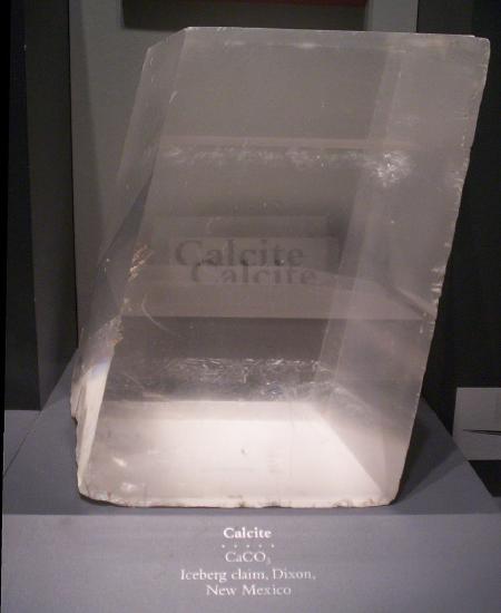 Transparent calcite crystal in the shape of a rhombohedron. The word 'calcite' is behind the specimen and appears twice through the mineral.