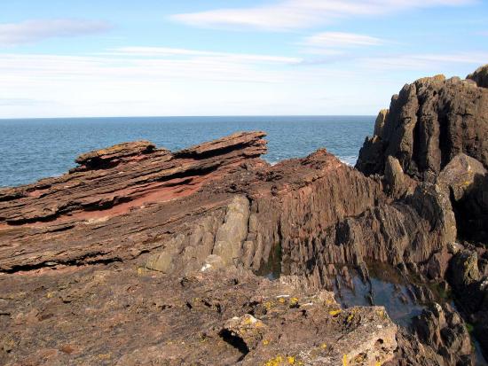 Brown rocks are layered and tilted along the ocean.