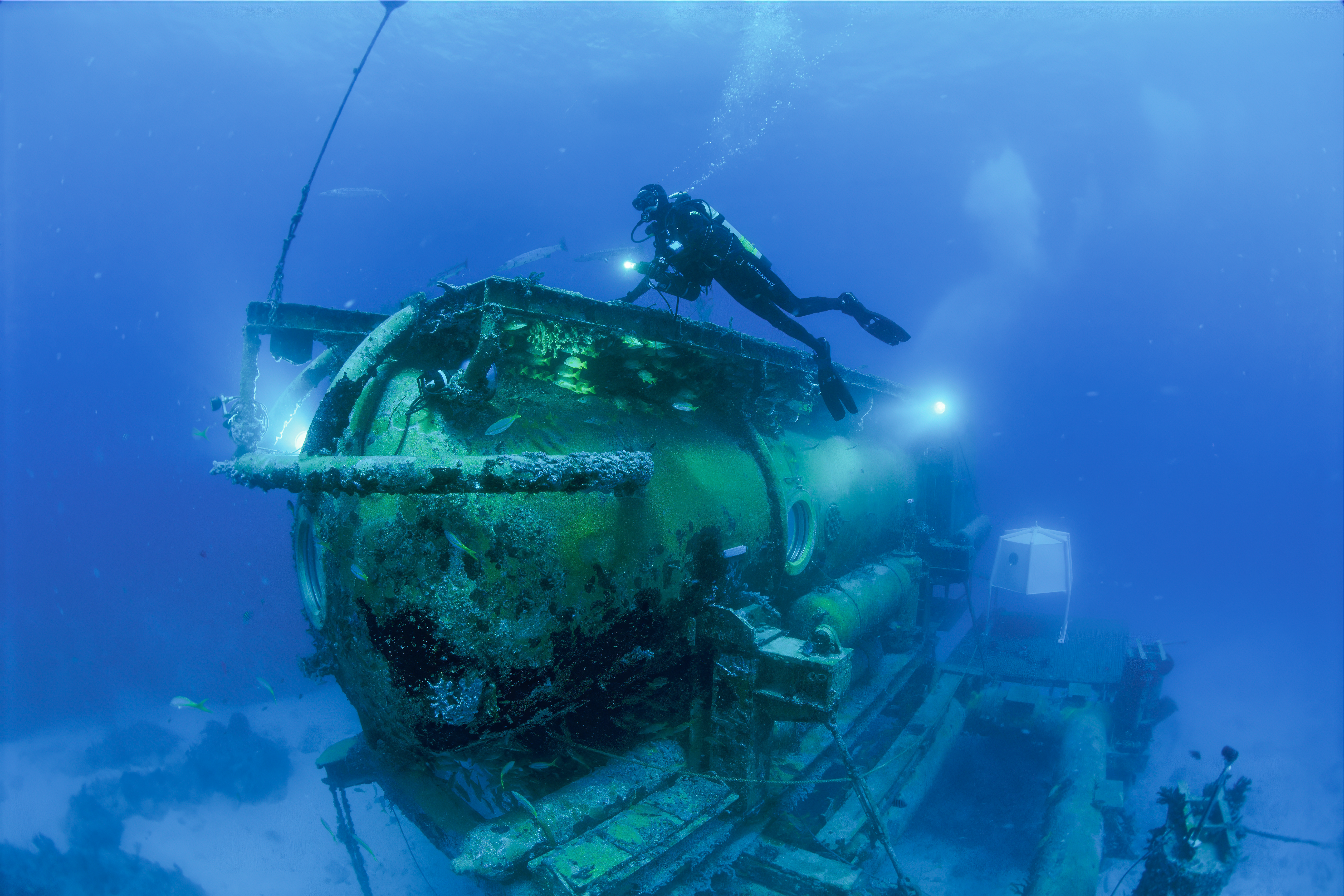 A scuba diver touching a large, cylindrical structure with lights and portholes that is mounted on a sled and sitting on the seafloor.