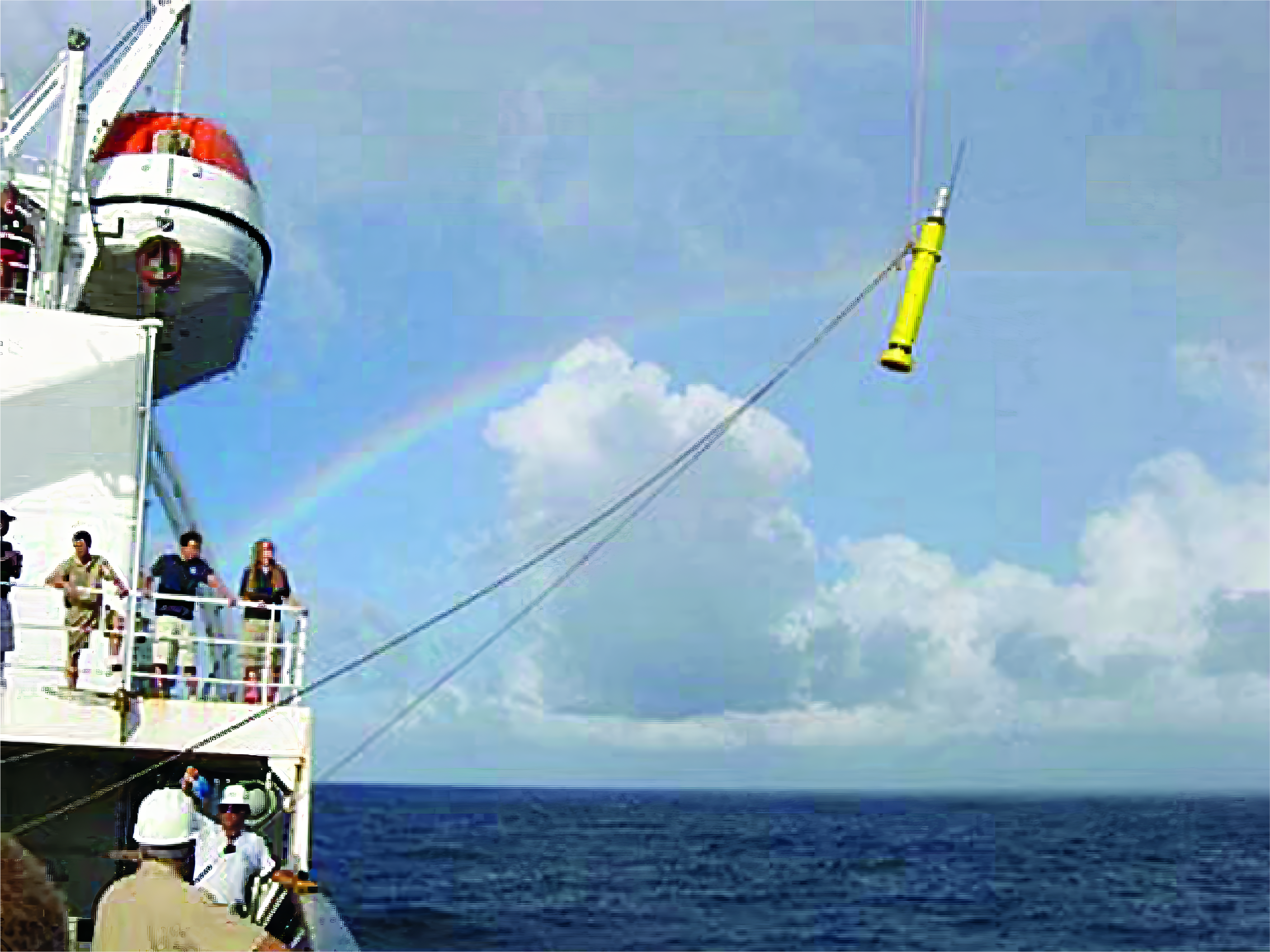 The side of a ship with many people watching a vertically oriented yellow cylinder being dropped into the ocean by a crane.