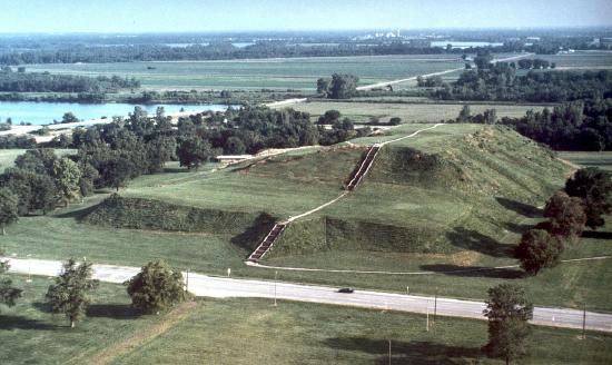 Aerial view of ancient earth mounds surrounded by greenery and a river in the background.