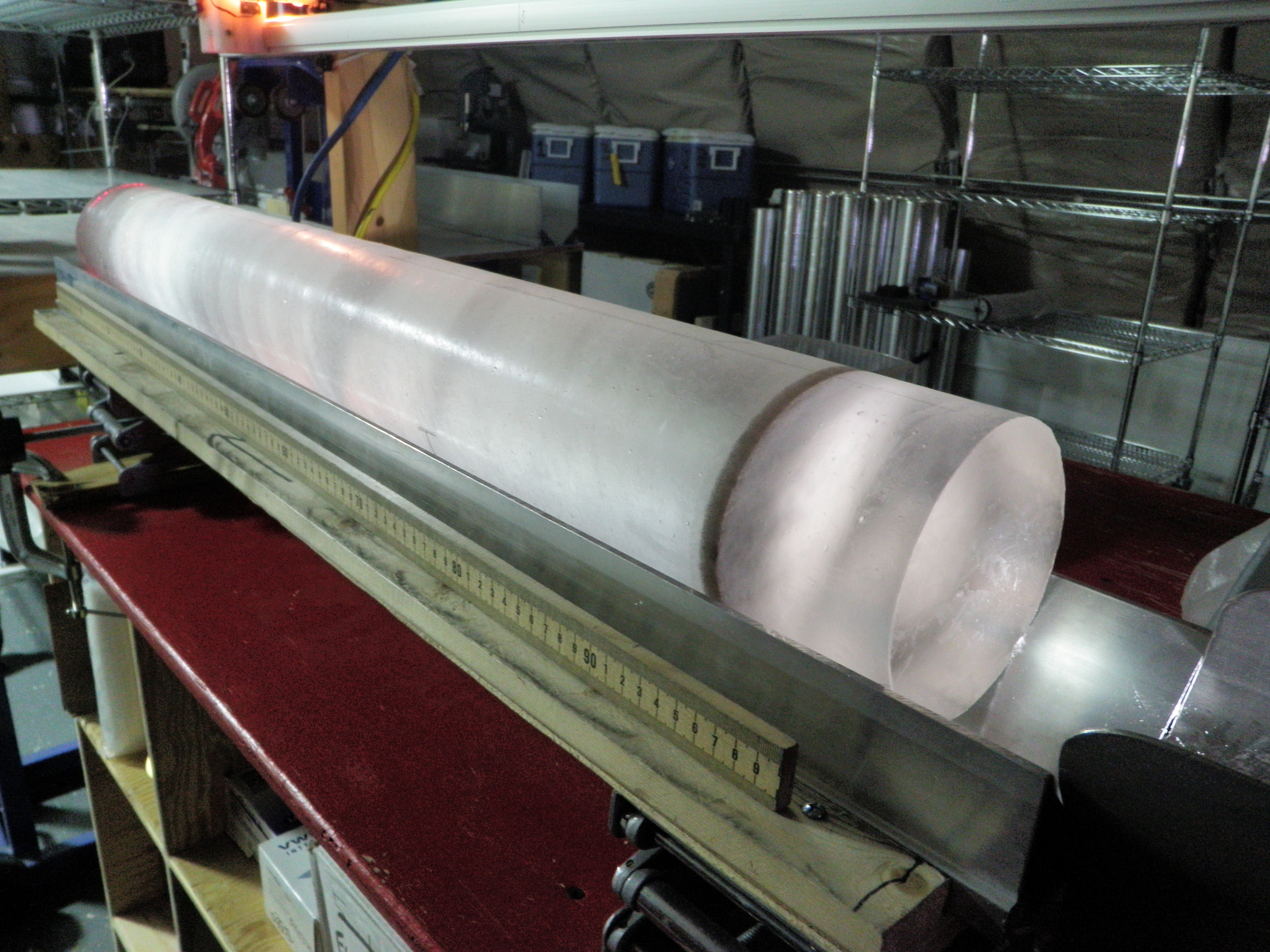 Large cylindrical ice core resting on a measuring cradle inside a cold laboratory processing facility. Details in Caption. 