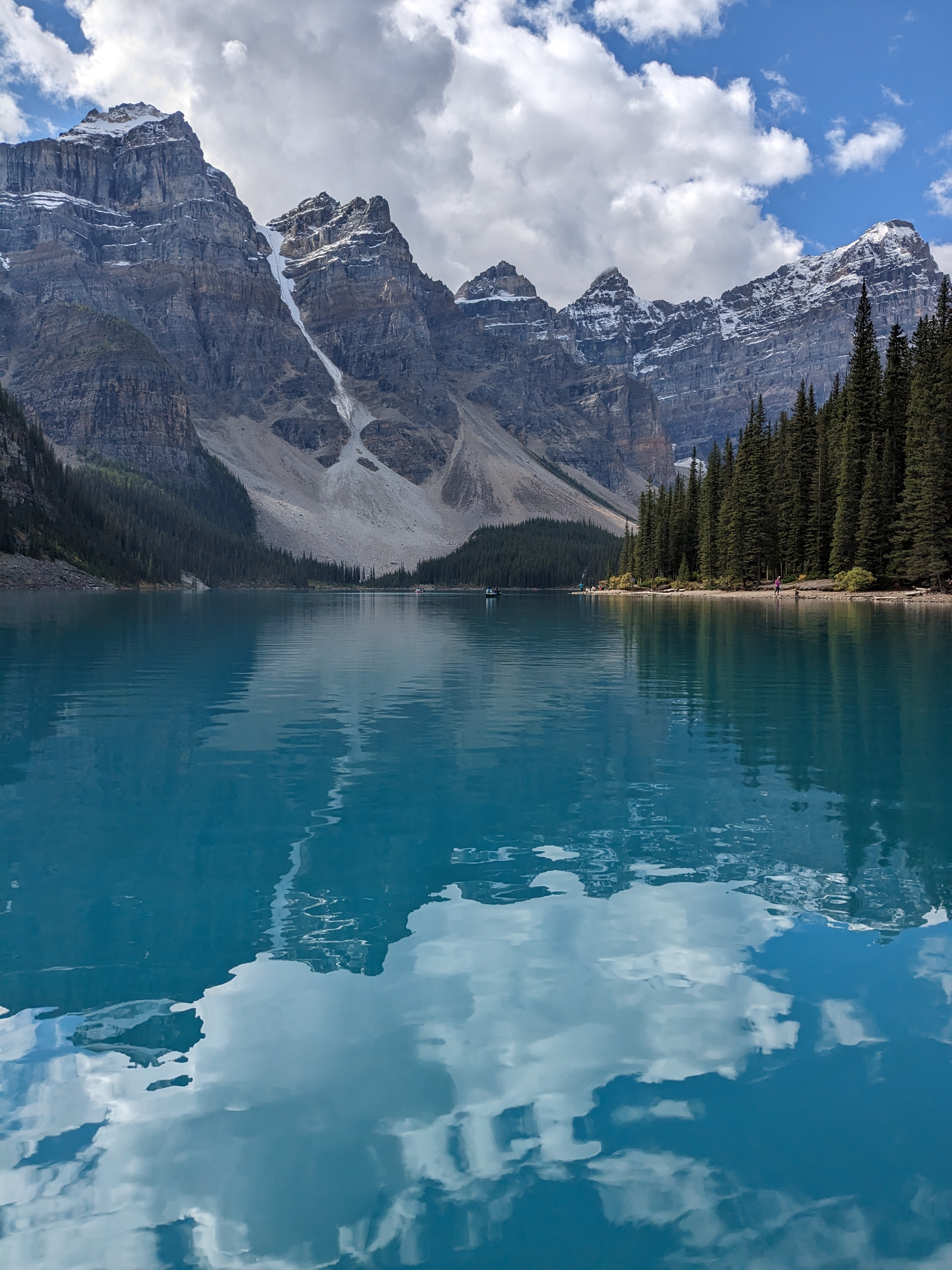 Turquoise alpine lake reflecting snow-dusted mountains and pine trees under a cloudy sky.