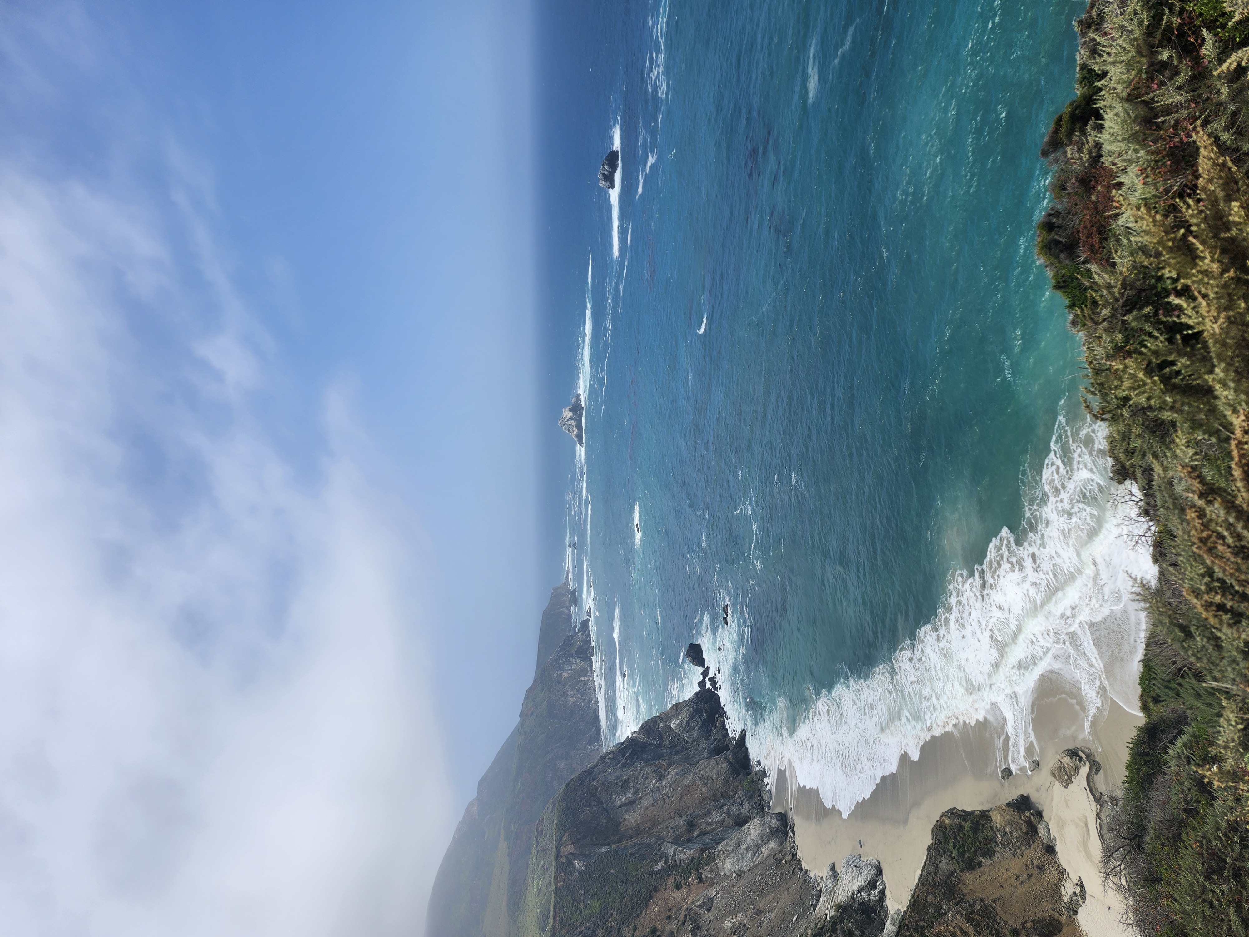 Steep rocky cliffs meet a vibrant blue ocean with white surf crashing onto a sandy beach below.