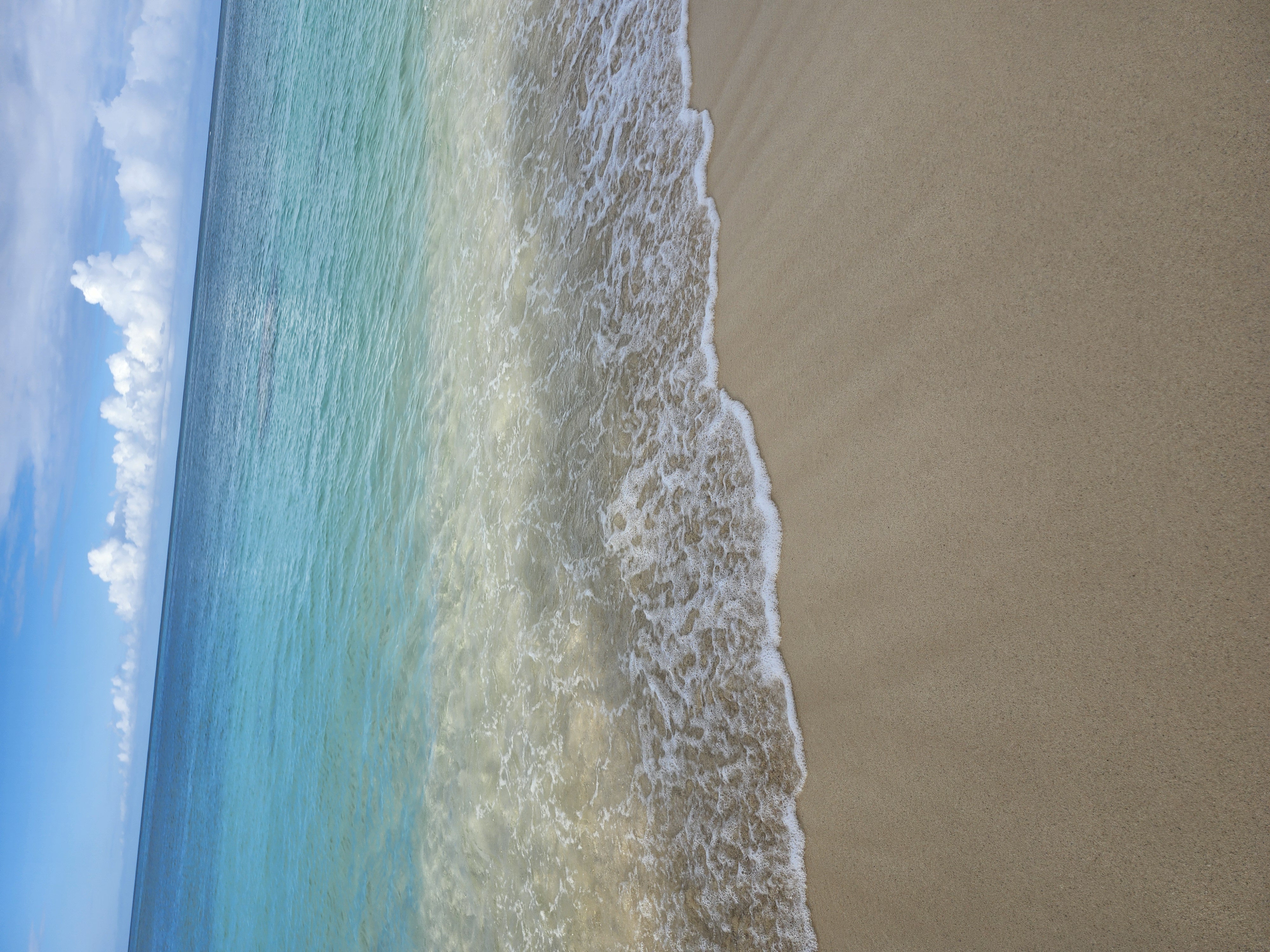 Clear turquoise ocean waves gently lapping onto a sandy beach under a blue sky with white clouds