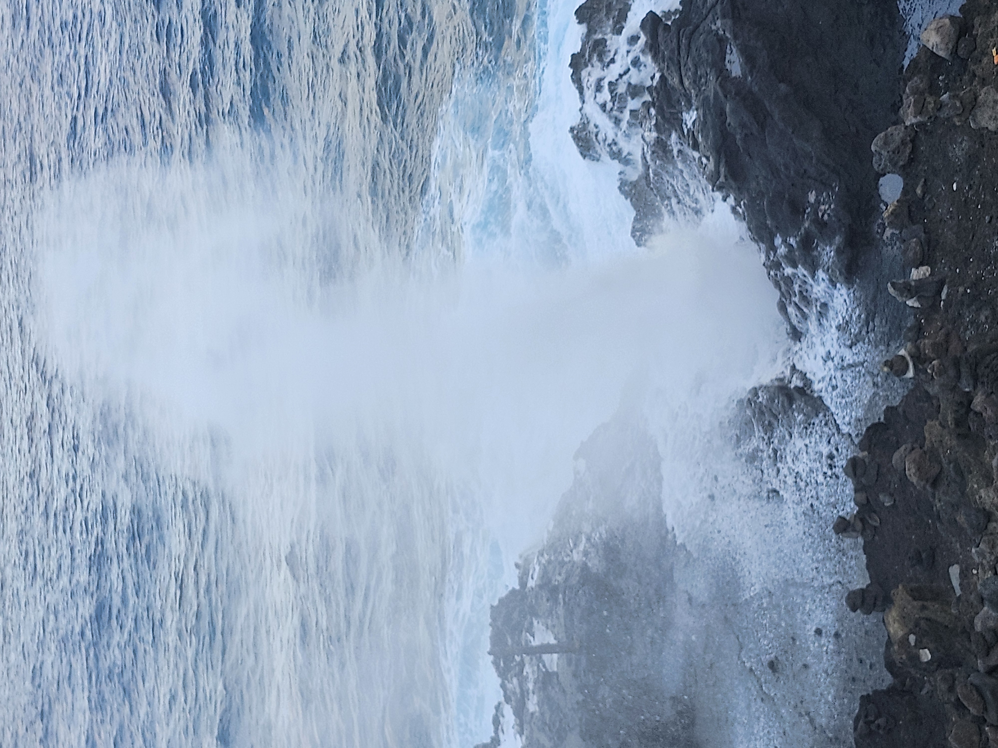 A tall plume of ocean water blasts upward out of a rocky blowhole along a volcanic coastline.