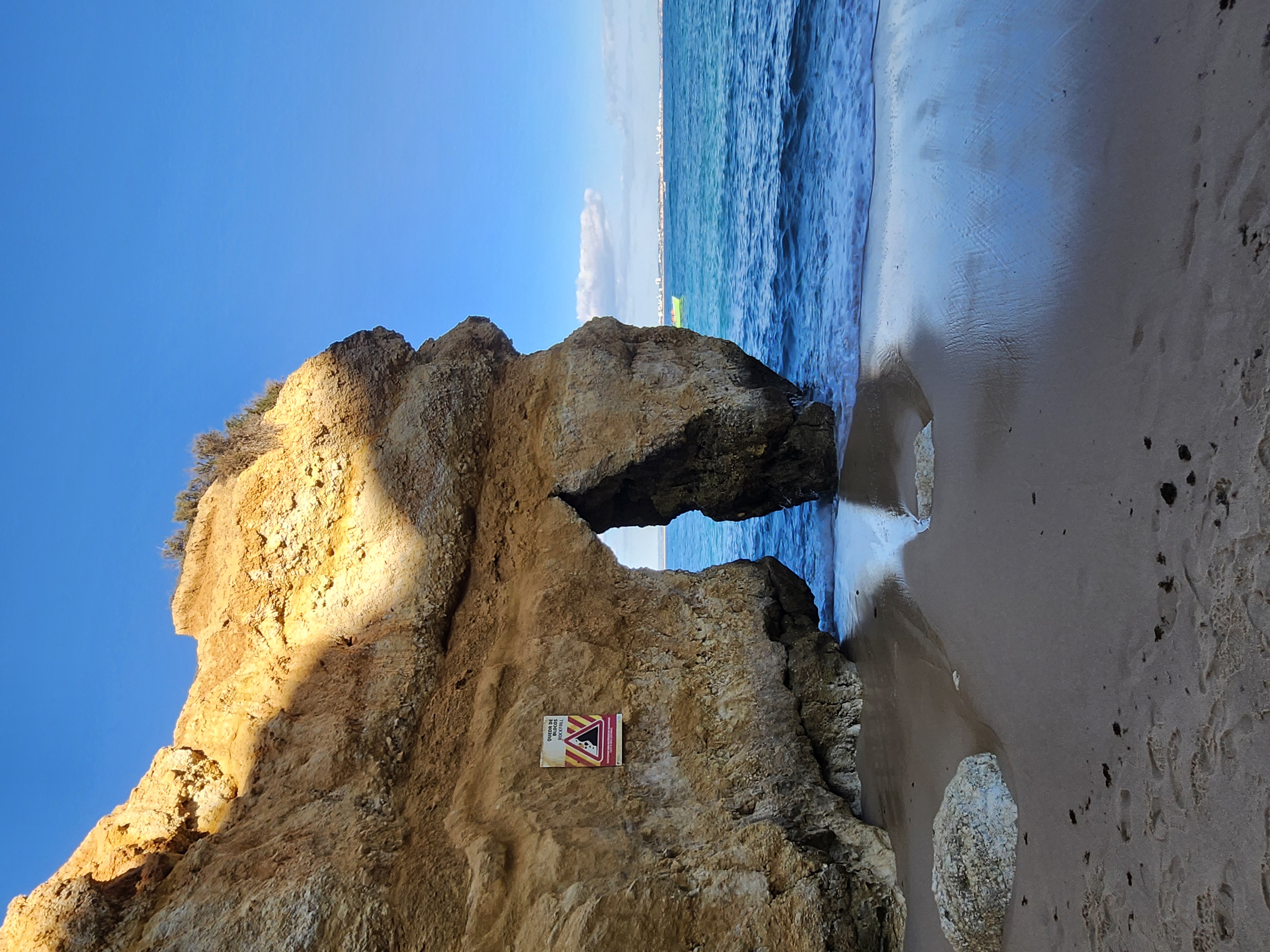 A natural limestone sea arch stands on a sandy beach next to the calm blue ocean under a clear sky.