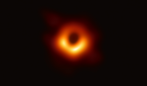 Blurry telescope photo of a fuzzy red halo around an entirely black center. The black center represents the first photograph of an actual black hole captured in 2019.